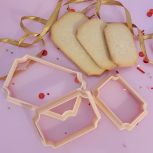 May include: Overhead shot of three baked cookies and three light pink cookie cutters in a tag shape. A gold ribbon and red confetti are scattered on a light purple surface.