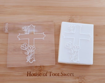 May include: A clear acrylic cookie stamp featuring a cross design with floral accents. The stamp is next to a cookie with the same embossed design. The text "House of Toot Sweet" is visible.