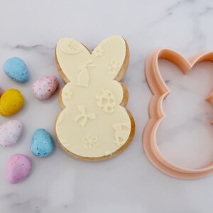 May include: An Easter-themed image featuring a bunny-shaped cookie with embossed designs of bunnies, eggs, and flowers. A matching cookie cutter sits beside the cookie. Colorful speckled candy eggs in shades of blue, pink, yellow, and green are scattered around.