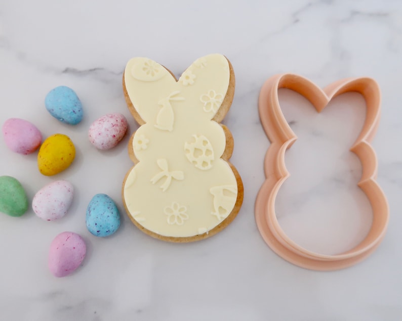May include: An Easter-themed image featuring a bunny-shaped cookie with embossed designs of bunnies, eggs, and flowers. A matching cookie cutter sits beside the cookie. Colorful speckled candy eggs in shades of blue, pink, yellow, and green are scattered around.