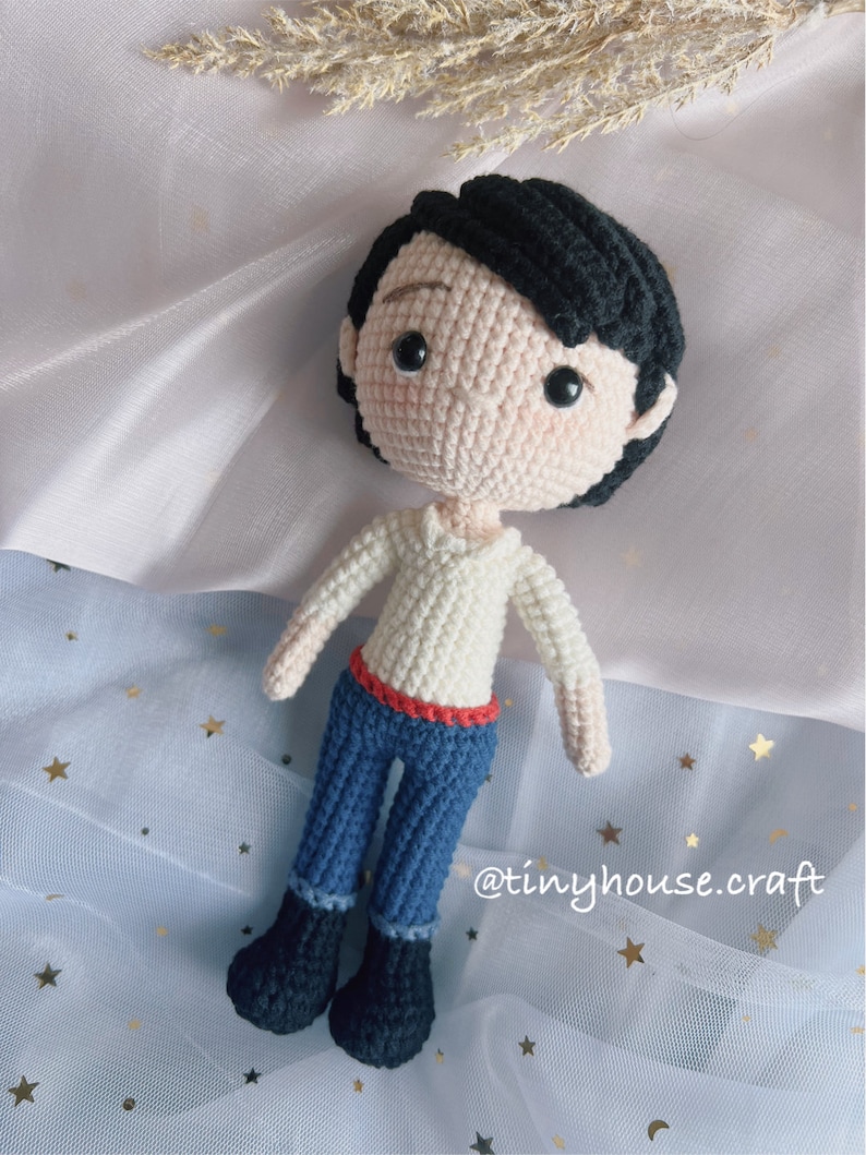 May include: Crocheted amigurumi doll with black hair, white shirt, red belt, blue jeans, and black shoes. The doll is on a light blue background with gold stars. The text "@tinyhouse.craft" is in the lower right corner of the image.