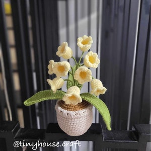 Puede incluir: Una planta de lirio de los valles de crochet en una maceta blanca con un borde marrón. La planta tiene flores de color amarillo claro y hojas verdes.