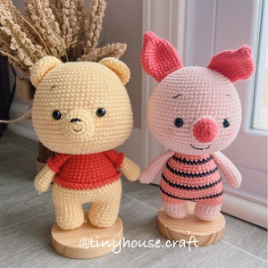 May include: Two crocheted stuffed animals, Winnie the Pooh and Piglet, standing on wooden bases. Winnie the Pooh is yellow with a red shirt and Piglet is pink with black and white stripes.