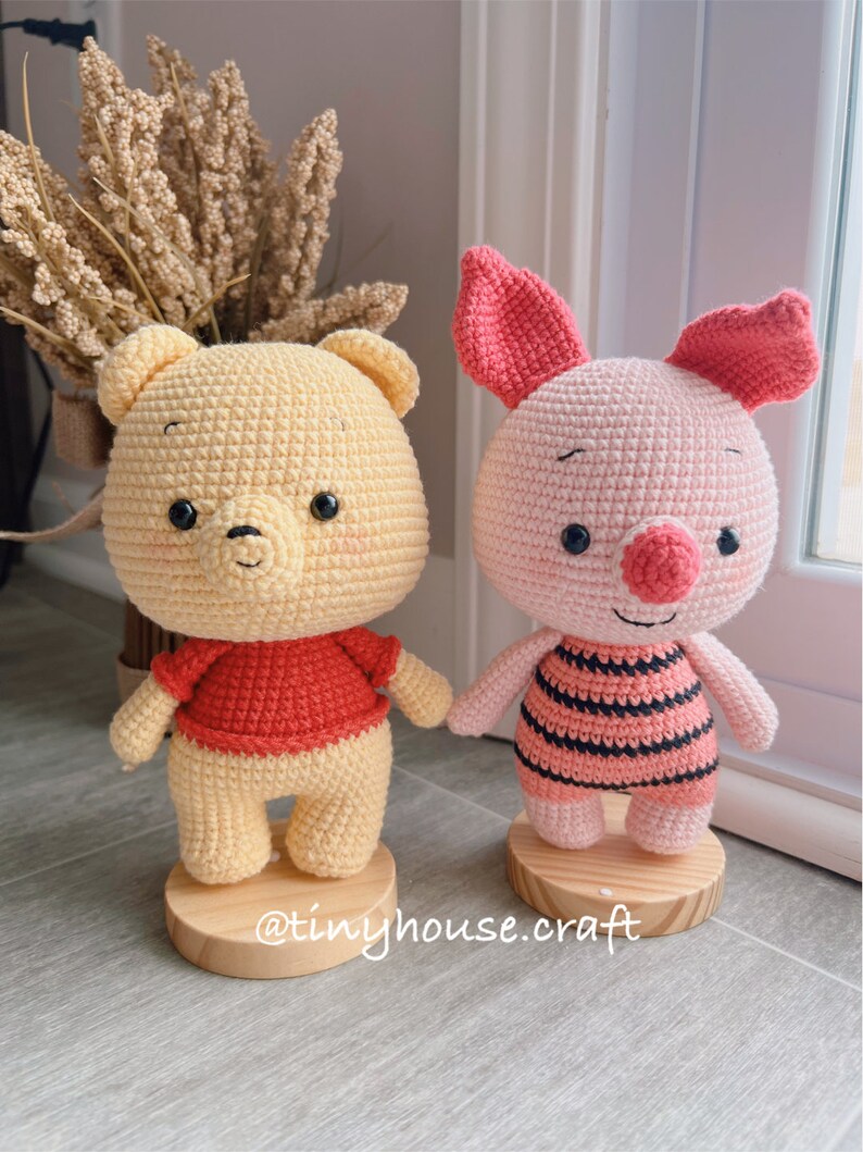 May include: Two crocheted stuffed animals, Winnie the Pooh and Piglet, standing on wooden bases. Winnie the Pooh is yellow with a red shirt and Piglet is pink with black and white stripes.