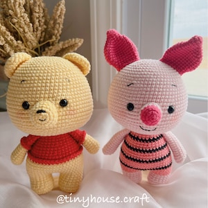 May include: Two crocheted stuffed animals, a yellow bear with a red shirt and a pink pig with black and pink striped clothing. The bear has a smiling face with black eyes and a small black nose. The pig has a smiling face with black eyes and a pink nose. The background is a white surface with a small bouquet of dried flowers.