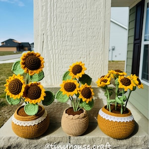 May include: Three crocheted sunflower plants in brown pots with white lace trim. The sunflowers are yellow with brown centers and green leaves. The pots are sitting on a brick surface. @tinyhouse.craft