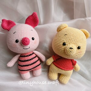 May include: Two crocheted stuffed animals, a pink pig and a yellow bear, are sitting on a white fabric background. The pig is wearing a pink and black striped shirt and the bear is wearing a red shirt. The text "@tinyhouse.craft" is visible in the bottom right corner of the image.
