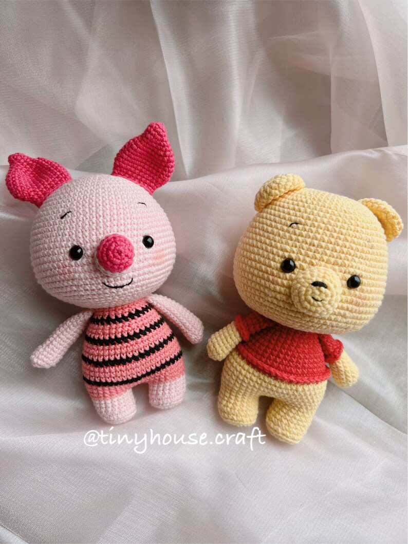 May include: Two crocheted stuffed animals, a pink pig and a yellow bear, are sitting on a white fabric background. The pig is wearing a pink and black striped shirt and the bear is wearing a red shirt. The text "@tinyhouse.craft" is visible in the bottom right corner of the image.