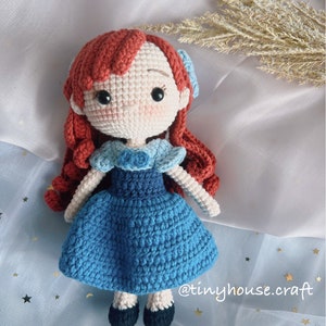 May include: A crocheted doll with red hair, a blue dress, and black shoes. The doll is wearing a blue bow in her hair. The doll is sitting on a blue and white fabric with gold stars.