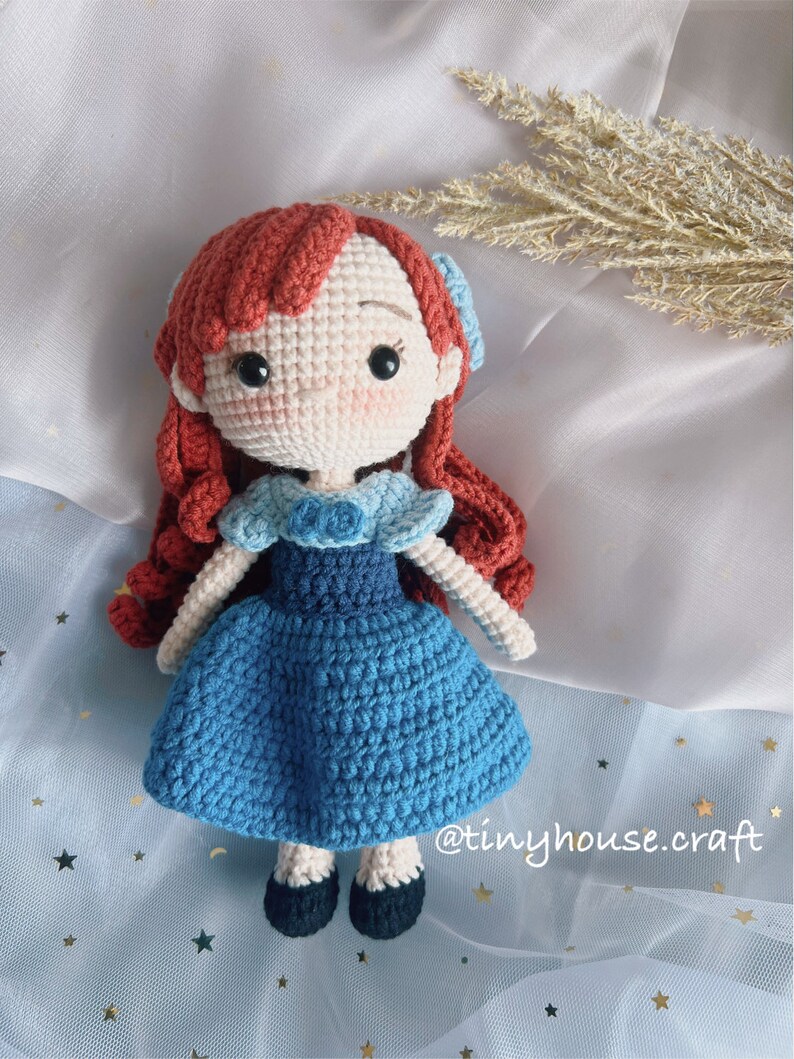 May include: A crocheted doll with red hair, a blue dress, and black shoes. The doll is wearing a blue bow in her hair. The doll is sitting on a blue and white fabric with gold stars.