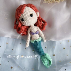 May include: Crocheted mermaid doll with red hair, a purple and white top, and a teal and green tail. The doll is lying on a blue fabric with gold stars.