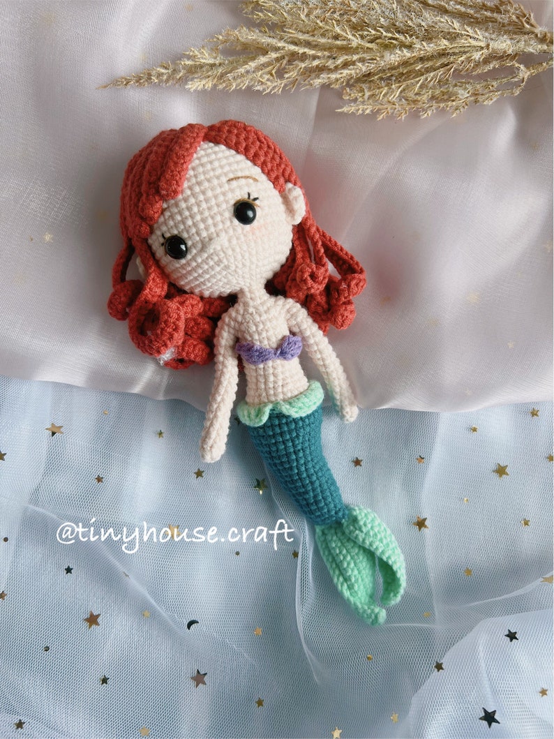May include: Crocheted mermaid doll with red hair, a purple and white top, and a teal and green tail. The doll is lying on a blue fabric with gold stars.