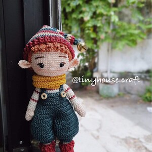 Op de afbeelding: Een gehaakte elfenpop met een rood-wit gestreepte muts, een gele sjaal en een blauwe overall. De pop heeft bruin haar en bruine ogen. De pop staat op een vensterbank. De tekst "@tinyhouse.craft" is zichtbaar op de achtergrond.