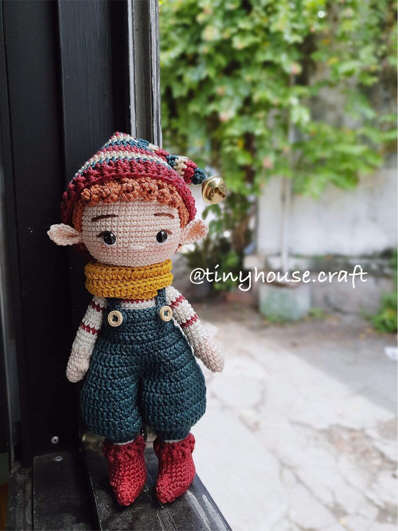 Op de afbeelding: Een gehaakte elfenpop met een rood-wit gestreepte muts, een gele sjaal en een blauwe overall. De pop heeft bruin haar en bruine ogen. De pop staat op een vensterbank. De tekst "@tinyhouse.craft" is zichtbaar op de achtergrond.
