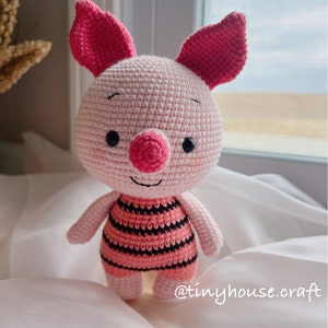 May include: A pink crocheted pig wearing a striped pink and black outfit. The pig has a pink nose and black eyes. The pig is standing on a white surface. @tinyhouse.craft