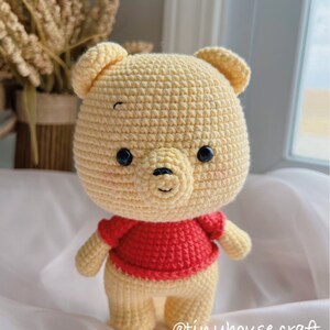 May include: A crocheted Winnie the Pooh plush toy with a red shirt. The toy is standing on a white surface. The text "@tinyhouse.craft" is visible in the bottom right corner of the image.