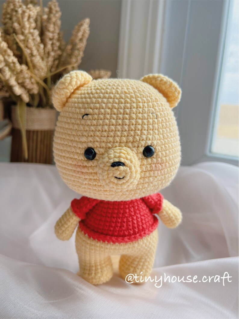 May include: A crocheted Winnie the Pooh plush toy with a red shirt. The toy is standing on a white surface. The text "@tinyhouse.craft" is visible in the bottom right corner of the image.