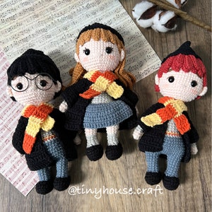 May include: Three crocheted dolls dressed as Harry Potter characters. The dolls are wearing black robes with yellow and orange scarves. The dolls are standing on a wooden surface.