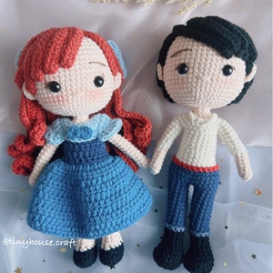 May include: Crocheted amigurumi dolls of Ariel and Prince Eric from the Disney movie The Little Mermaid. Ariel is wearing a blue dress with a white bodice and red hair. Prince Eric is wearing a white shirt, blue pants, and black shoes. The dolls are on a light blue background with gold stars.