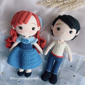 May include: Crocheted amigurumi dolls of Ariel and Prince Eric from the Disney movie The Little Mermaid. Ariel is wearing a blue dress with a red hair bow. Prince Eric is wearing a white shirt and blue pants. The dolls are sitting on a blue and white background with gold stars.