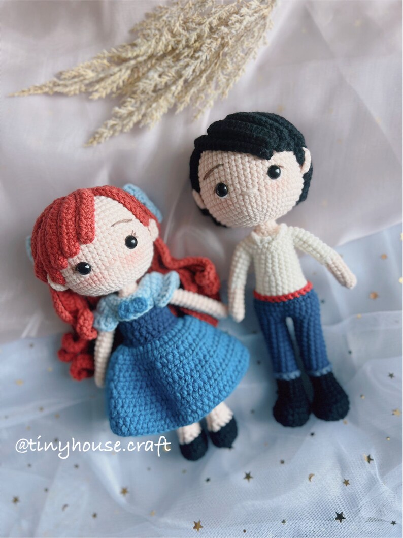 May include: Crocheted amigurumi dolls of Ariel and Prince Eric from the Disney movie The Little Mermaid. Ariel is wearing a blue dress with a white collar and red hair. Prince Eric is wearing a white shirt, blue pants, and black hair.