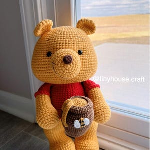 May include: A handmade crochet Winnie the Pooh plush toy. The bear is yellow with a red shirt and holds a brown honey pot with a white and yellow bee. The text "@tinyhouse.craft" is visible.