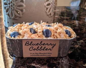 May include: A rectangular aluminum tin candle topped with textured tan wax crumbles and dark blue wax blueberries, featuring a label that reads Blueberry Cobbler, 16 oz. Soy Blended Wax, and cabinsidecandles.com, with three visible candle wicks.