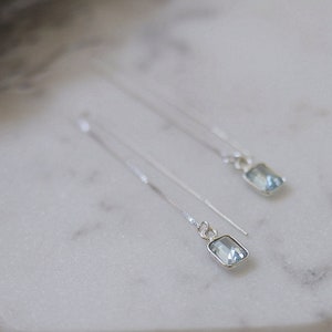 Könnte beinhalten: Ein Paar filigrane Silber-Ohrringe mit rechteckigen, hellblauen Edelsteinen. Die Ohrringe werden auf einer weißen, sechseckigen Oberfläche präsentiert, die ihr elegantes Design und ihren subtilen Glanz hervorhebt.
