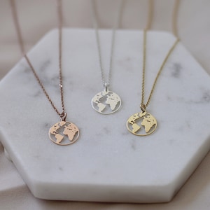 Könnte beinhalten: Drei zarte Halsketten mit Globus-Anhängern in Roségold, Silber und Gold. Jeder Anhänger hat ein ausgeschnittenes Design der Weltkarte. Die Halsketten werden auf einer weißen, sechseckigen Oberfläche präsentiert.