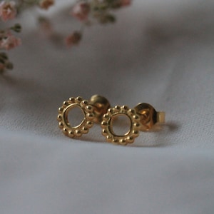 Könnte beinhalten: Ein Paar goldfarbene Ohrstecker mit floralem Design. Die Ohrringe haben eine kreisförmige Form mit kleinen, perlenbesetzten Details am Rand. Zarte rosa Blüten sind im Hintergrund zu sehen.