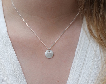 May include: A delicate silver necklace featuring a small, textured, circular pendant. The pendant hangs from a fine chain, creating a minimalist and elegant look. The necklace is shown against a white background.