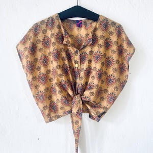 Pode incluir: Uma blusa bege de manga curta com um padrão repetido de desenhos florais vermelhos e azuis. A blusa tem gola, fecho de botões e uma gravata na cintura. A blusa está pendurada em um cabide preto contra uma parede branca.