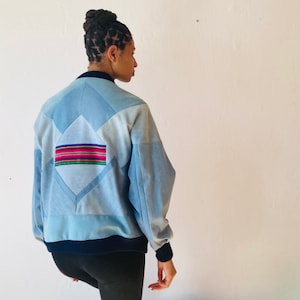 Könnte beinhalten: Eine hellblaue Bomberjacke mit geometrischem Patchwork-Design. Die Jacke hat einen dunkelblauen gerippten Kragen, Bündchen und Bund. Ein farbenfroher Streifen-Patch ist auf der Rückseite aufgenäht.