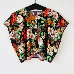 Könnte beinhalten: Ein kurzärmliges, geknöpftes Oberteil mit einem lebendigen Blumenmuster. Der schwarze Stoff ist mit einer Mischung aus roten, orangefarbenen, weißen und beigen Blumen mit grünen Blättern bedeckt. Das Oberteil hängt an einem schwarzen Kleiderbügel.