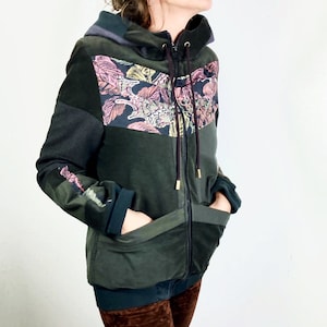 Könnte beinhalten: Ein dunkelgrüner Kapuzenpullover mit einem Blumendesign auf der Vorderseite. Der Pullover hat einen Reißverschluss und zwei Taschen.