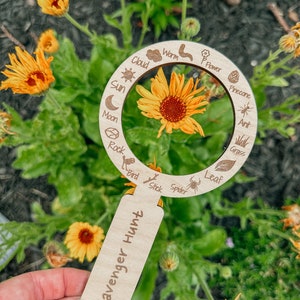May include: A wooden magnifying glass with a scavenger hunt label. The magnifying glass has a circle with various nature items listed around the edge, including a sun, moon, cloud, flower, worm, rock, bird, stick, spider, leaf, pine cone, ant, and grass. The magnifying glass is held in front of a yellow flower.
