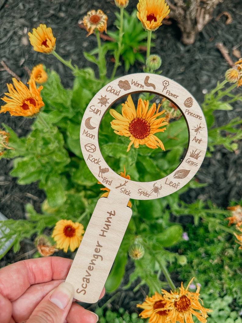 May include: A wooden magnifying glass with a scavenger hunt label. The magnifying glass has a circle with various nature items listed around the edge, including a sun, moon, cloud, flower, worm, rock, bird, stick, spider, leaf, pine cone, ant, and grass. The magnifying glass is held in front of a yellow flower.