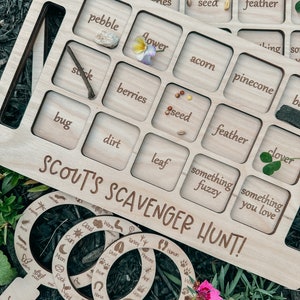 May include: Wooden board game with a grid of squares, each with a word and a space for a natural object. The words include "pebble", "flower", "acorn", "pinecone", "stick", "berries", "seed", "feather", "bug", "dirt", "leaf", "something fuzzy", and "something you love". The board is labeled "Scout's Scavenger Hunt!" and includes magnifying glass-shaped cutouts with nature-themed words.