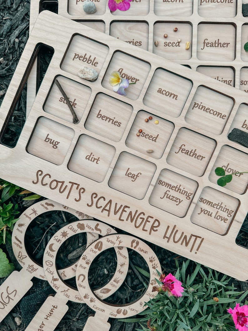 May include: Wooden board game with a grid of squares, each with a word and a space for a natural object. The words include "pebble", "flower", "acorn", "pinecone", "stick", "berries", "seed", "feather", "bug", "dirt", "leaf", "something fuzzy", and "something you love". The board is labeled "Scout's Scavenger Hunt!" and includes magnifying glass-shaped cutouts with nature-themed words.