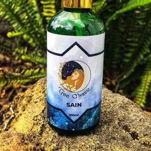 May include: A green glass bottle with a gold cap and a blue and white label. The label features a woman with a crown of leaves and the text "Tree Mystic" and "SAIN". The bottle contains 100 ml of liquid.