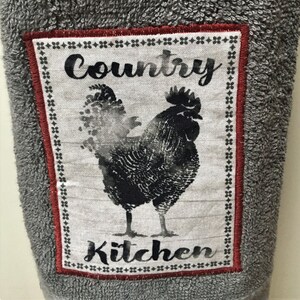 May include: A gray kitchen towel with a red and white embroidered patch featuring a black rooster and the text "Country Kitchen".