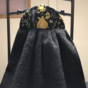 May include: Black kitchen towel with a yellow and black pineapple design on the top. The towel is hanging on a black metal rack.