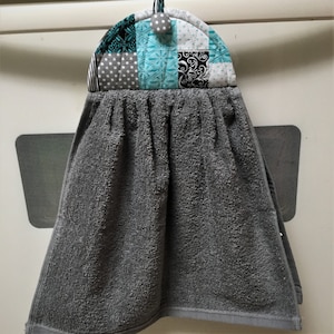 May include: Gray kitchen towel with a patchwork design hanging on a hook. The patchwork design features black, white, gray, and teal fabrics.
