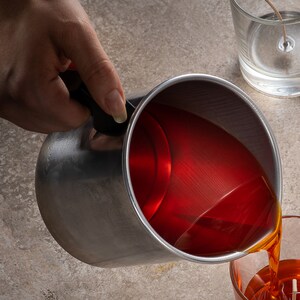 May include: A person pouring red wax from a silver pitcher into a clear glass jar. The wax is a deep red color and is being poured in a smooth stream.