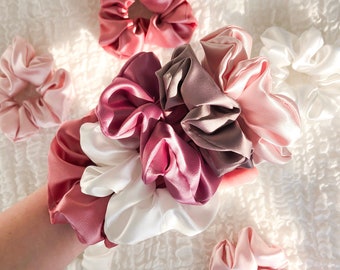 May include: A collection of satin scrunchies in various shades of pink, white, and taupe. The scrunchies are gathered and ruffled, with a soft, shiny texture. The scrunchies are arranged on a white, textured surface.