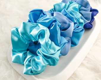 May include: A collection of satin scrunchies in various shades of blue, from light turquoise to deep indigo, arranged on a white, curved tray. The scrunchies have a shiny, smooth texture, suggesting a soft and luxurious feel. The background is a fluffy, white surface.