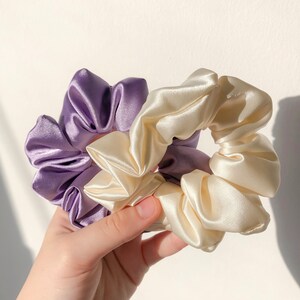 May include: Two satin scrunchies, one purple and one cream, are held in a hand. The scrunchies are made of soft, silky fabric and are perfect for holding hair in place.