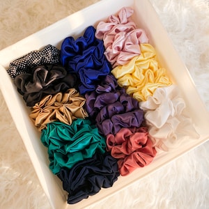 May include: A white box filled with a variety of colorful scrunchies. The scrunchies are made of satin and come in a variety of colors, including pink, blue, green, and yellow.