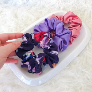 May include: Three hair scrunchies in a white ceramic dish. One scrunchie is purple, one is pink, and one is black with a floral print.