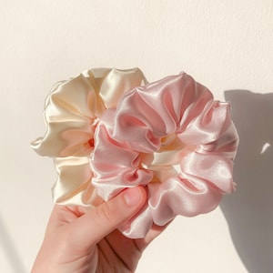 May include: Two satin scrunchies, one in a light pink and one in a cream color. The scrunchies are tied together and held in a hand.